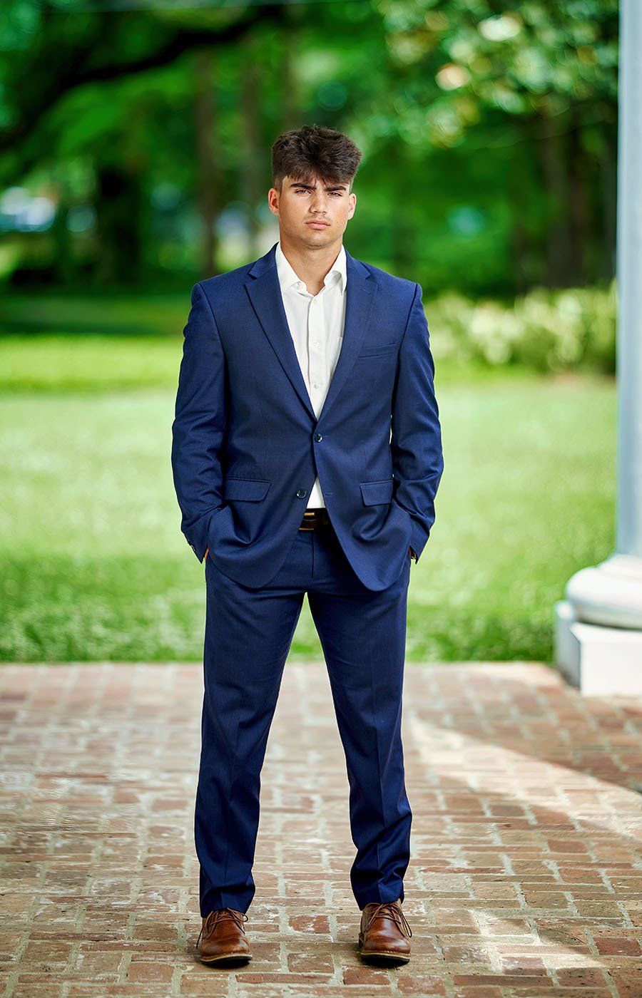 A young man in a blue suit is standing on a brick sidewalk with his hands in his pockets.