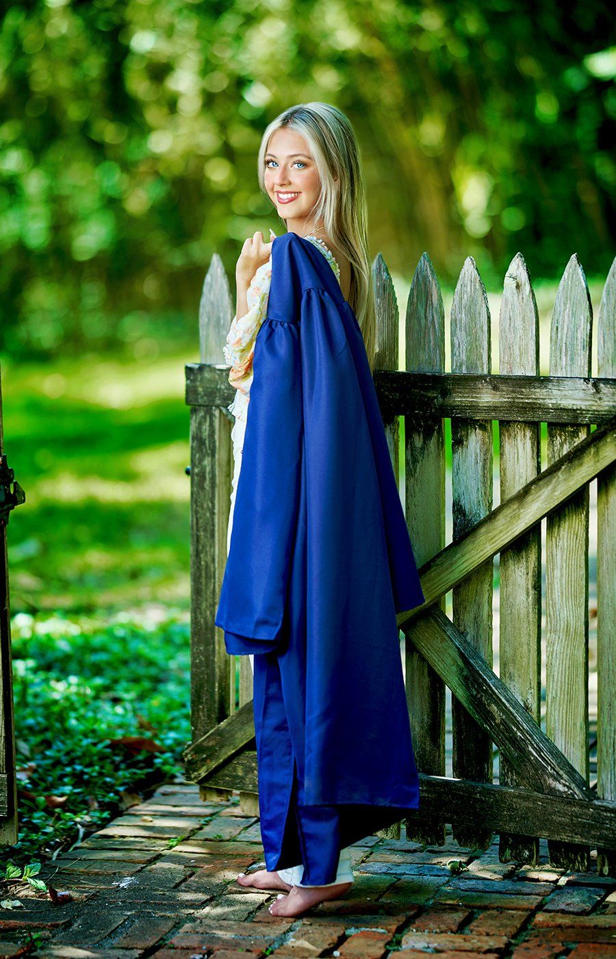 A woman in a blue graduation cap and gown is standing next to a wooden fence.