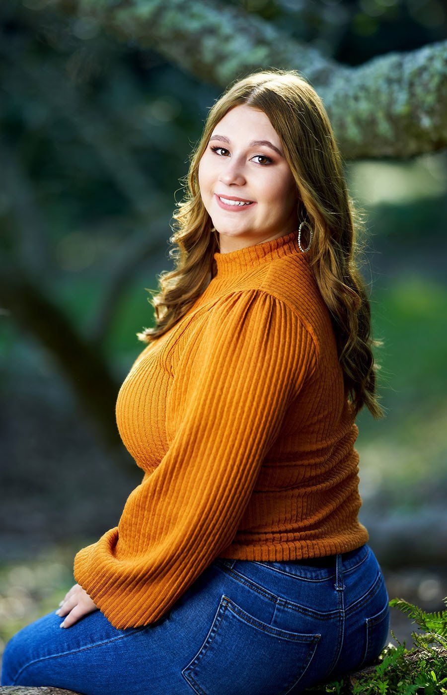 A woman in a yellow sweater and blue jeans is sitting under a tree.