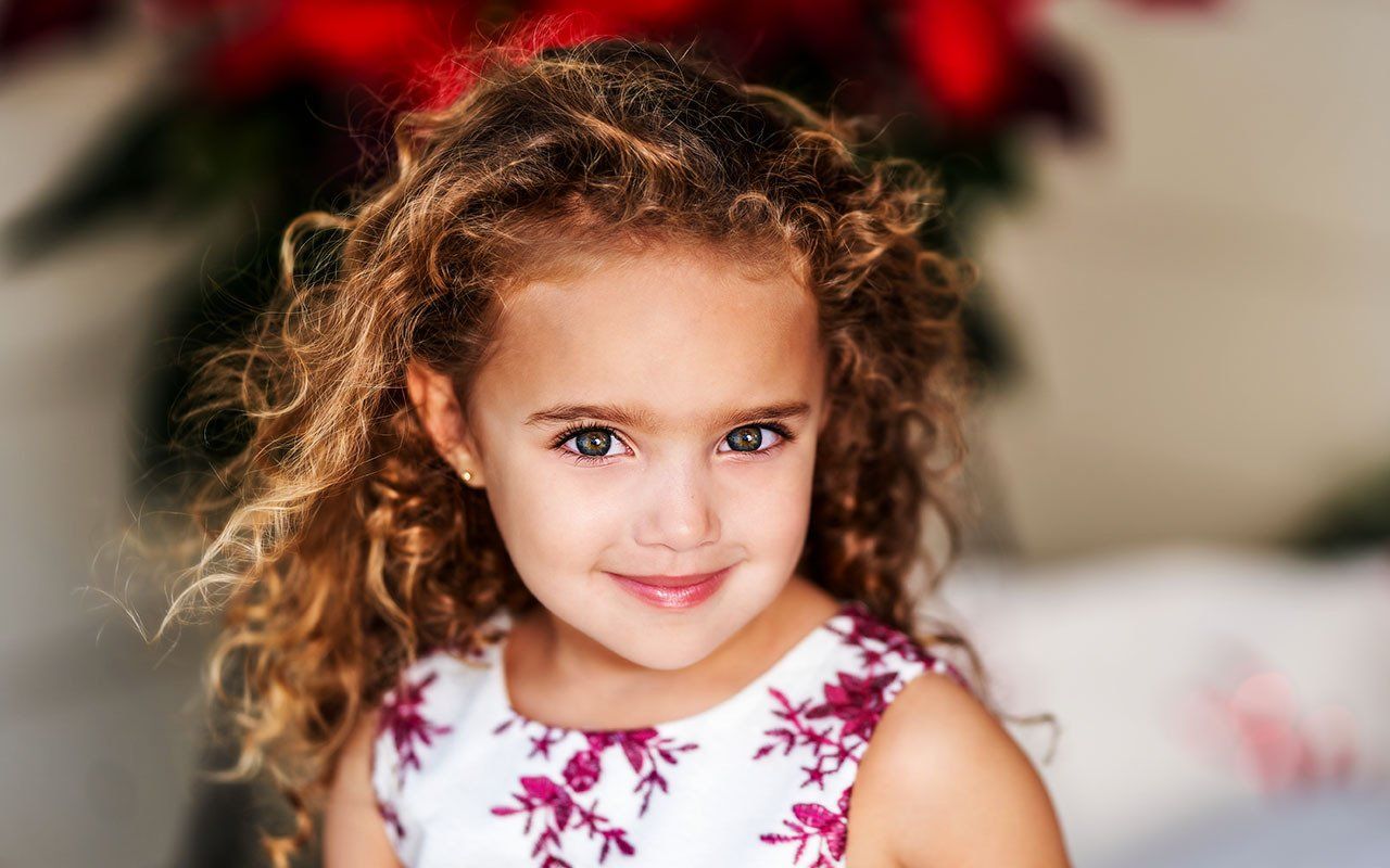 A little girl with curly hair is smiling in front of a poinsettia.
