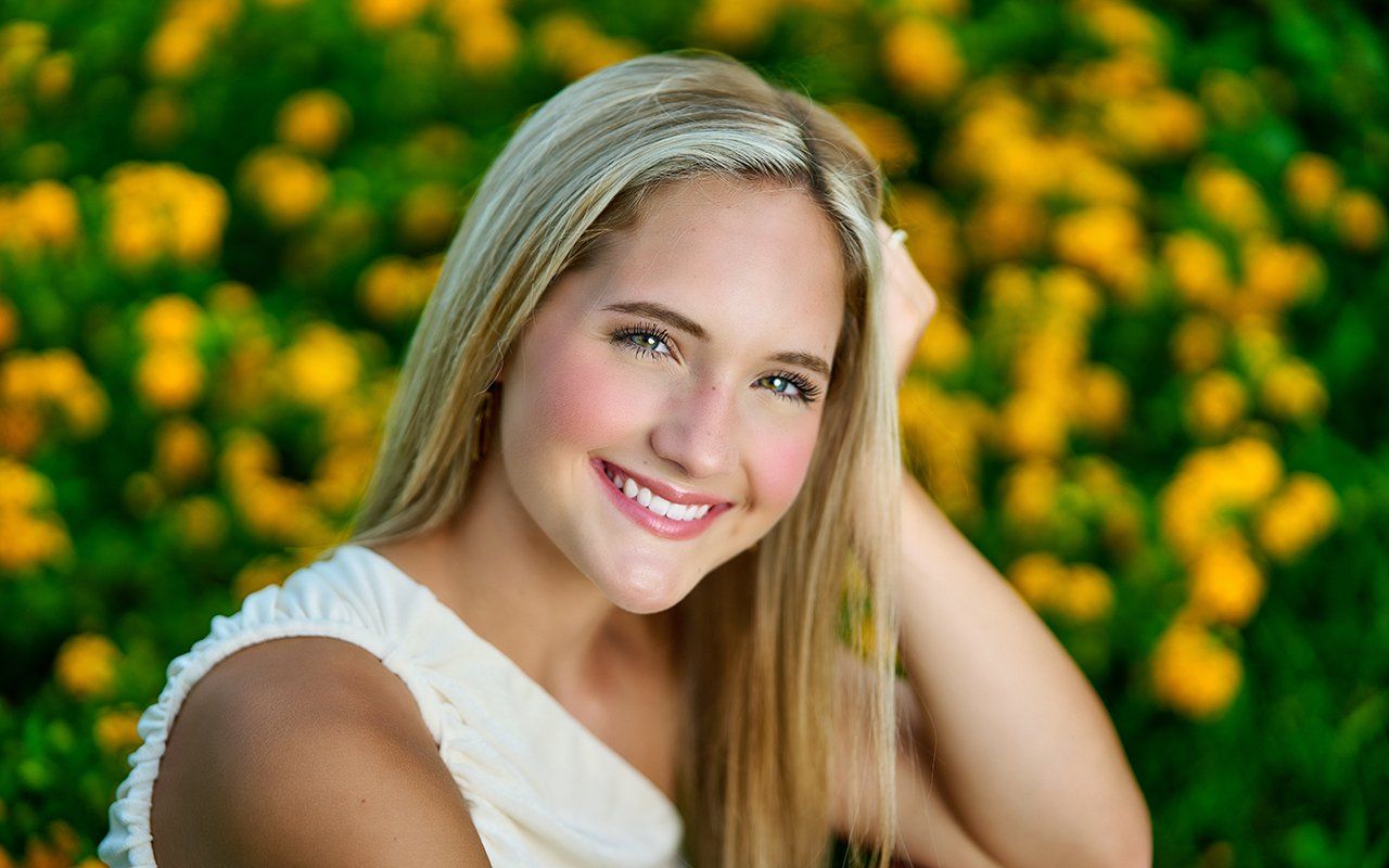 A young woman is smiling in front of a field of yellow flowers.