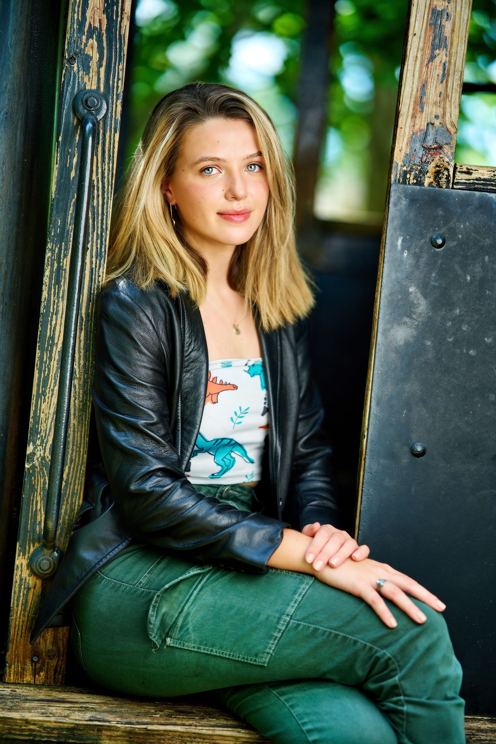 A woman in a leather jacket and green pants is sitting on a wooden bench.