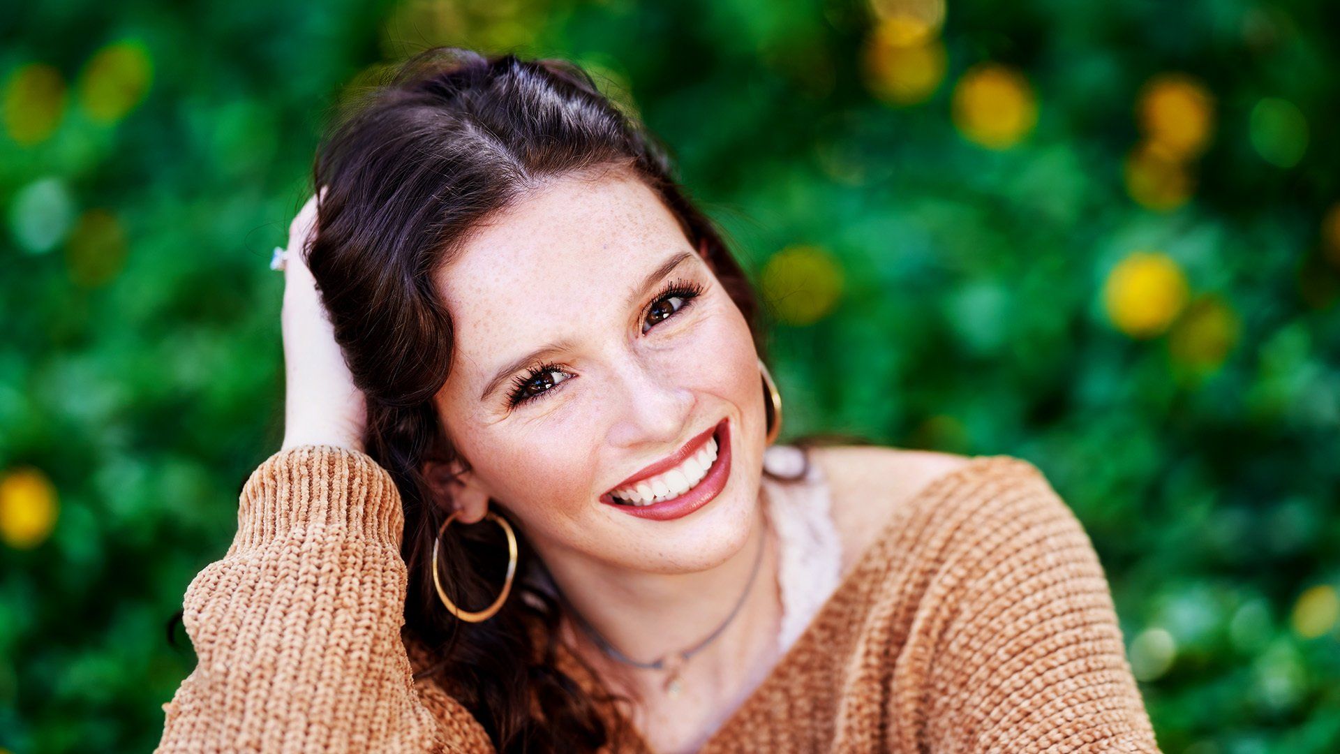 A young woman wearing a brown sweater and hoop earrings is smiling.