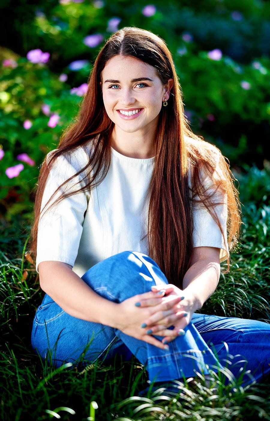 A young woman is sitting in the grass with her legs crossed and smiling.
