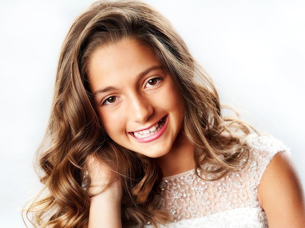 A young girl with long brown hair is wearing a white dress and smiling.