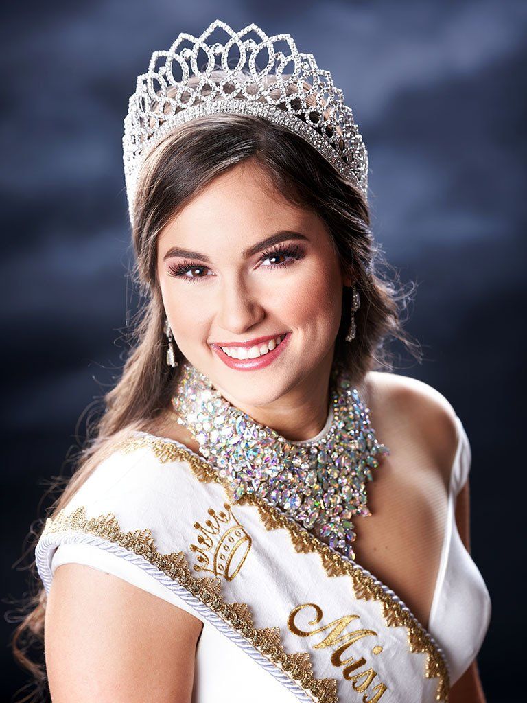 A woman wearing a tiara and a sash is smiling for the camera.