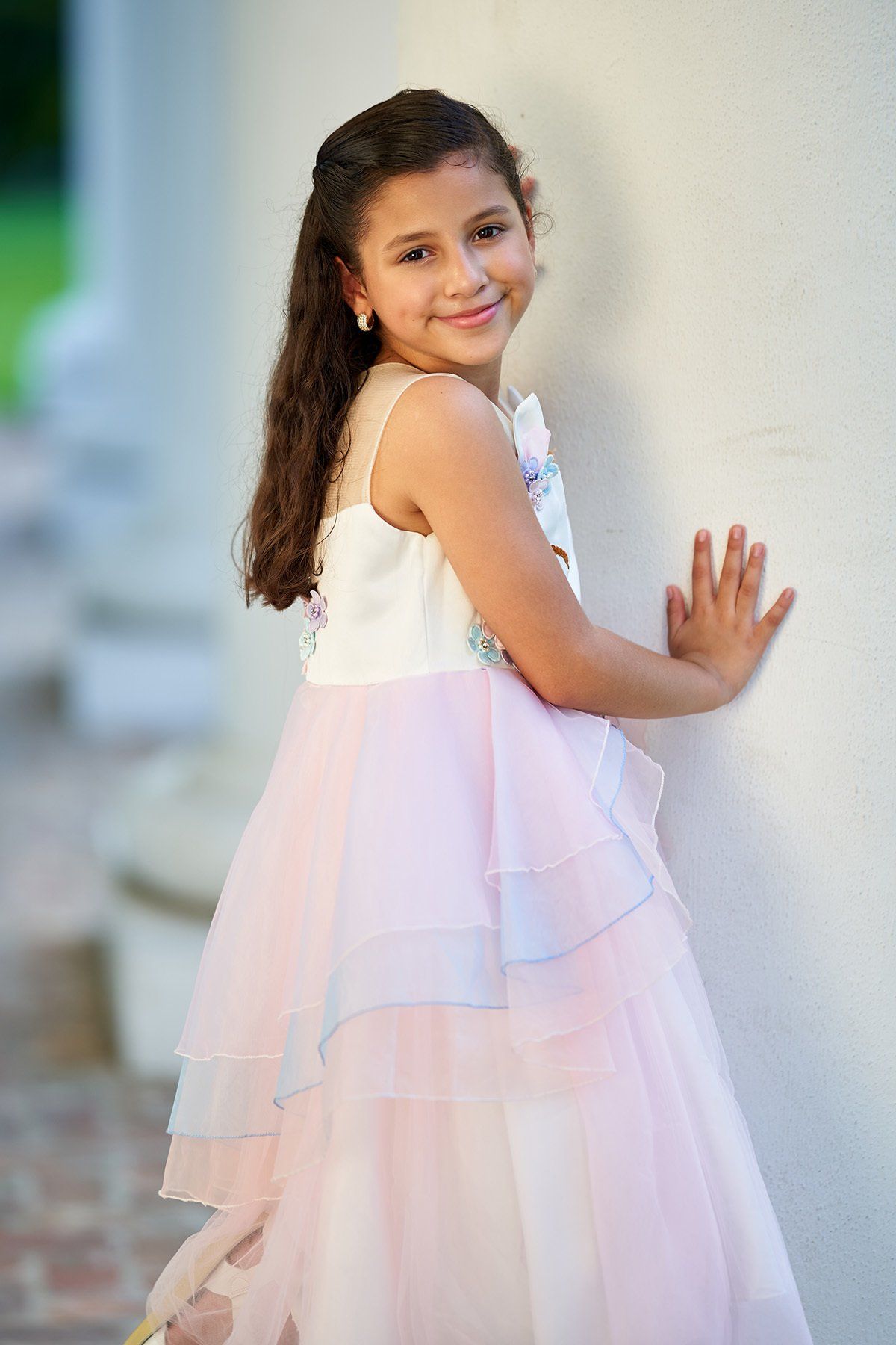 A little girl in a pink and white dress is leaning against a wall.