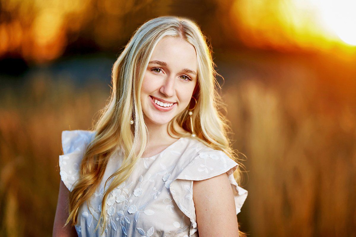 A young woman in a white dress is smiling in a field at sunset.
