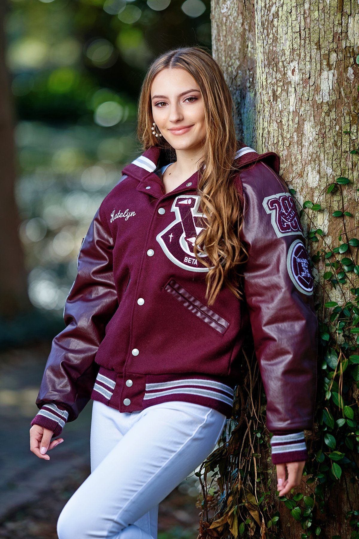 A young woman in a maroon letterman jacket is leaning against a tree.