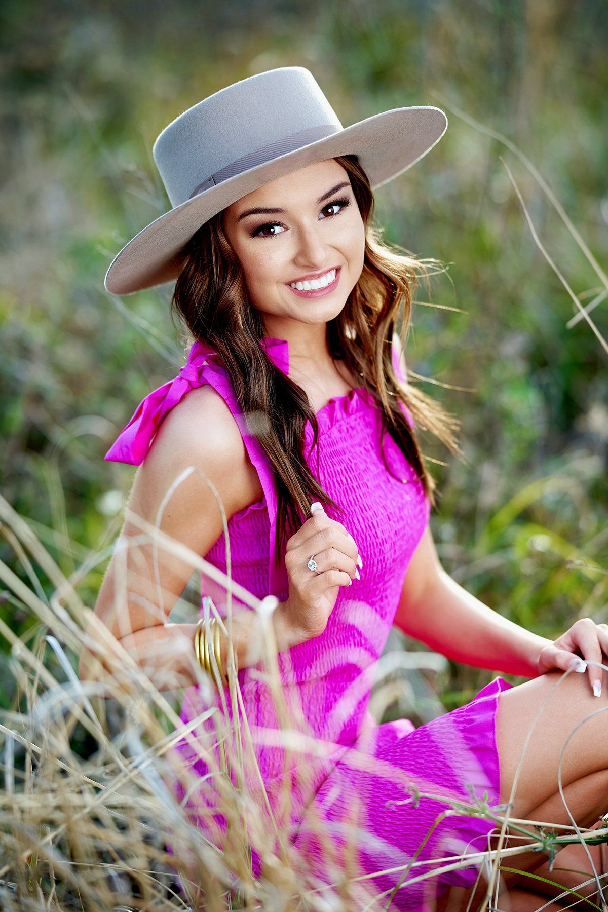 A woman in a pink dress and hat is sitting in the grass.