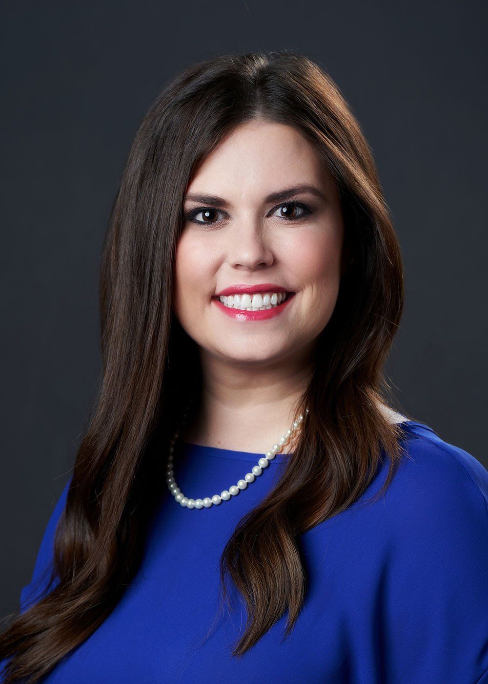 A woman wearing a blue shirt and a pearl necklace is smiling.