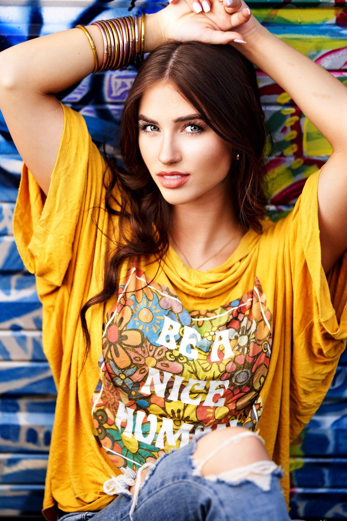 A woman wearing a yellow shirt that says be a nice human is standing in front of a graffiti wall.