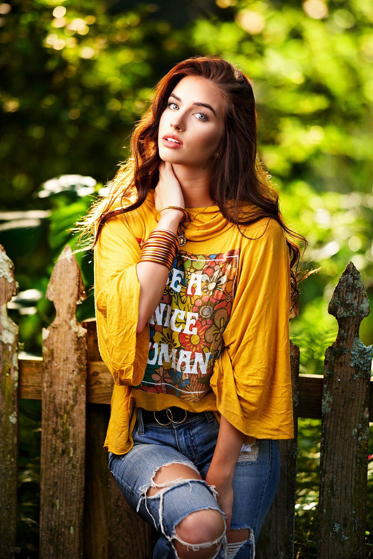 A woman in a yellow shirt and ripped jeans is sitting on a wooden fence.