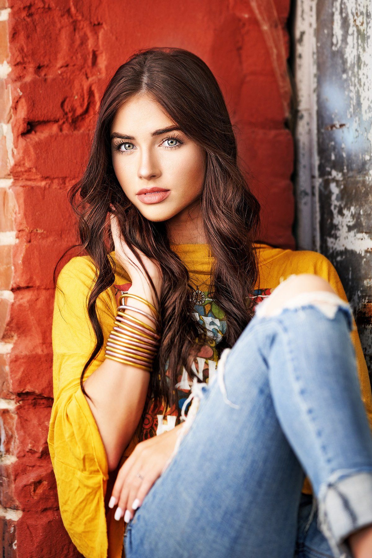 A woman in a yellow shirt and blue jeans is sitting in front of a brick wall.