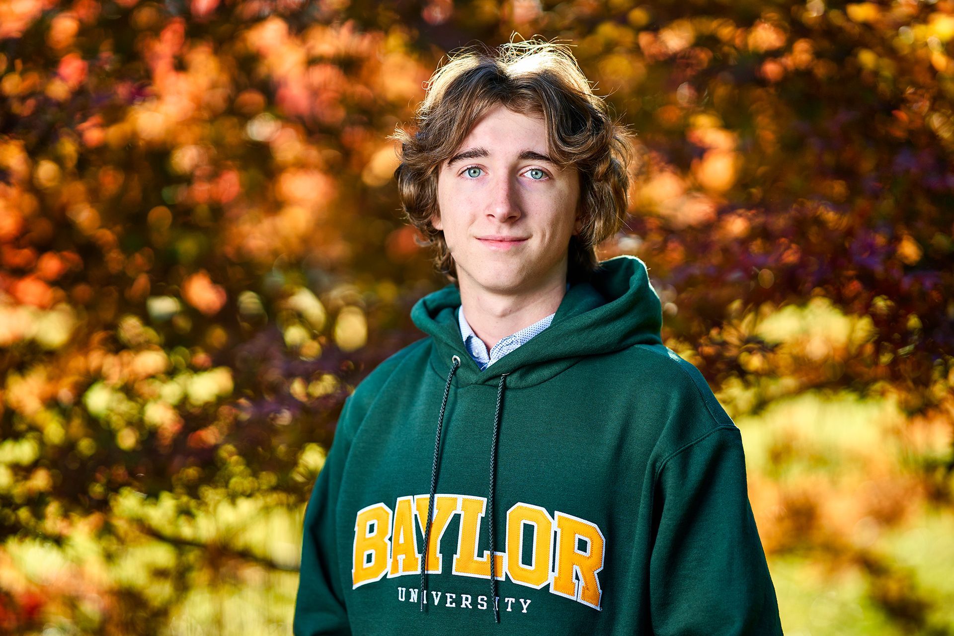 A young man wearing a green hoodie is standing in front of a tree.