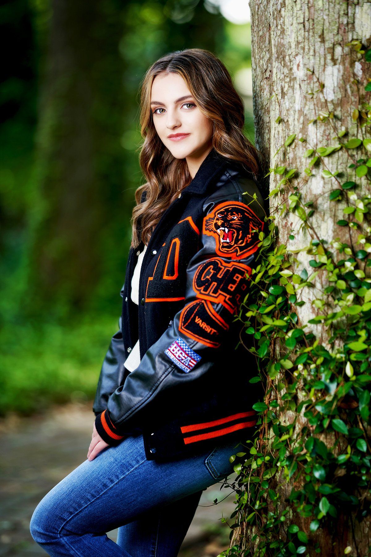 A woman in a letterman jacket is leaning against a tree.