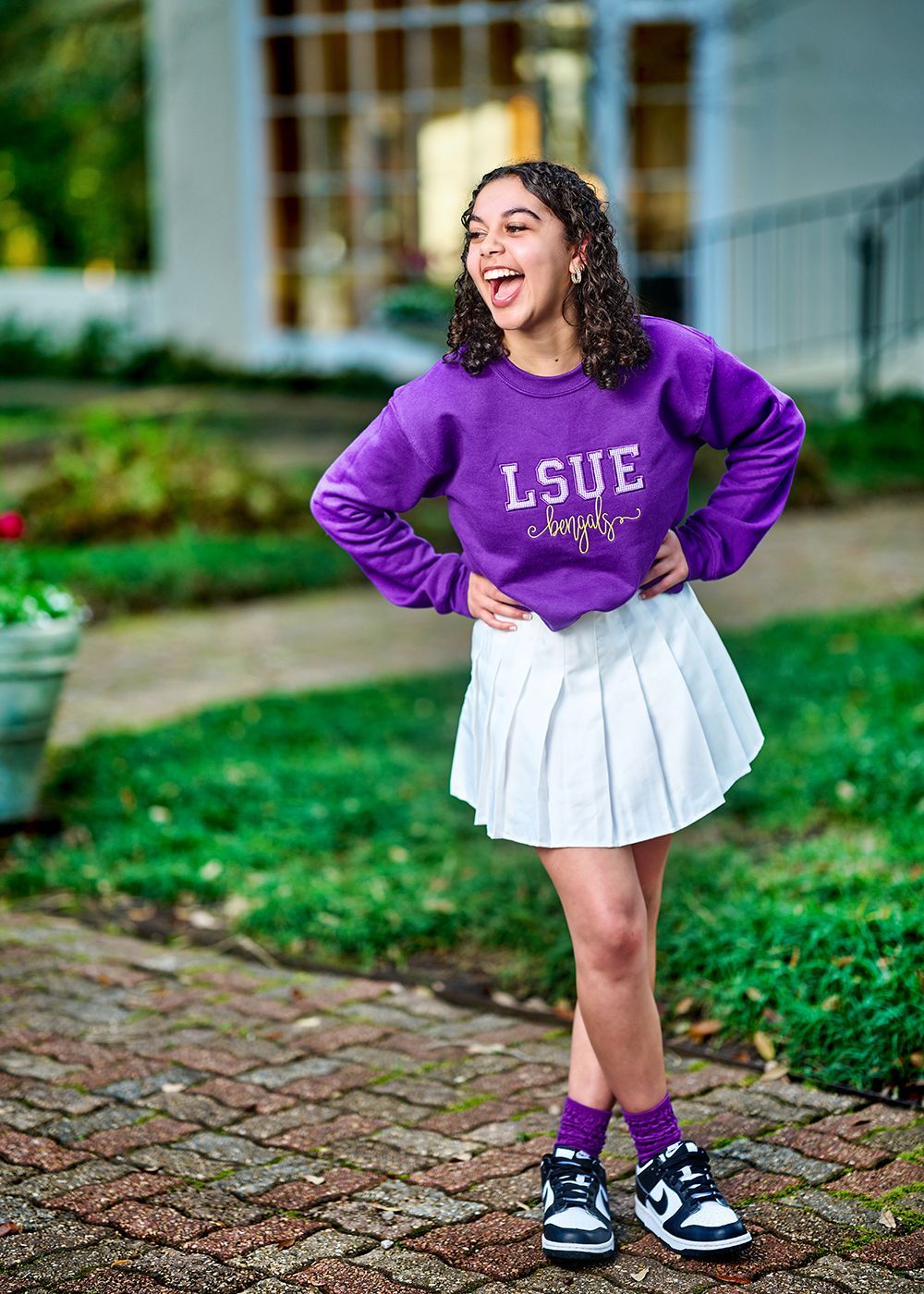 A young girl wearing a purple sweatshirt and a white pleated skirt is standing on a sidewalk.