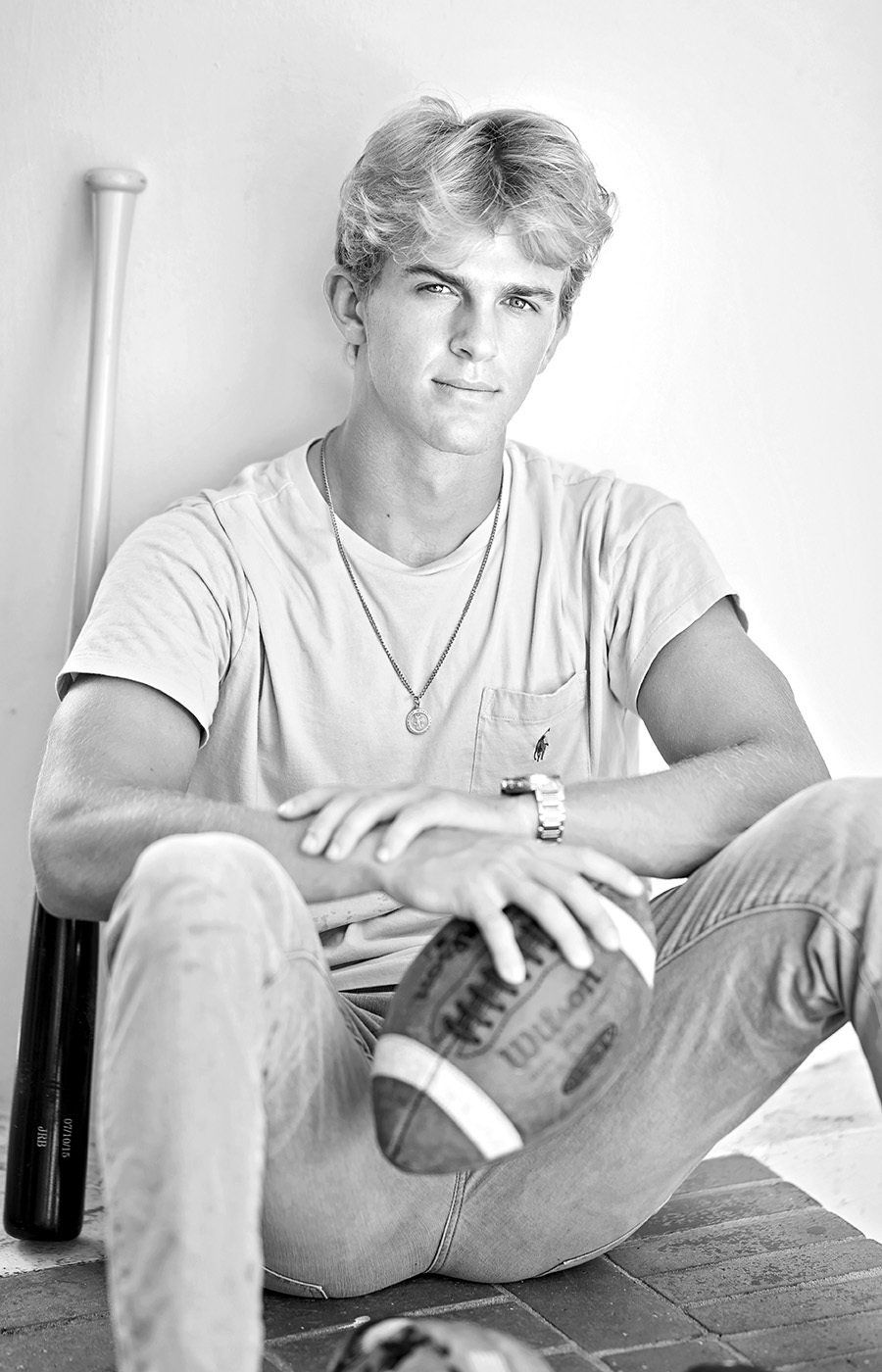 A young man is sitting on the floor holding a football and a baseball bat.