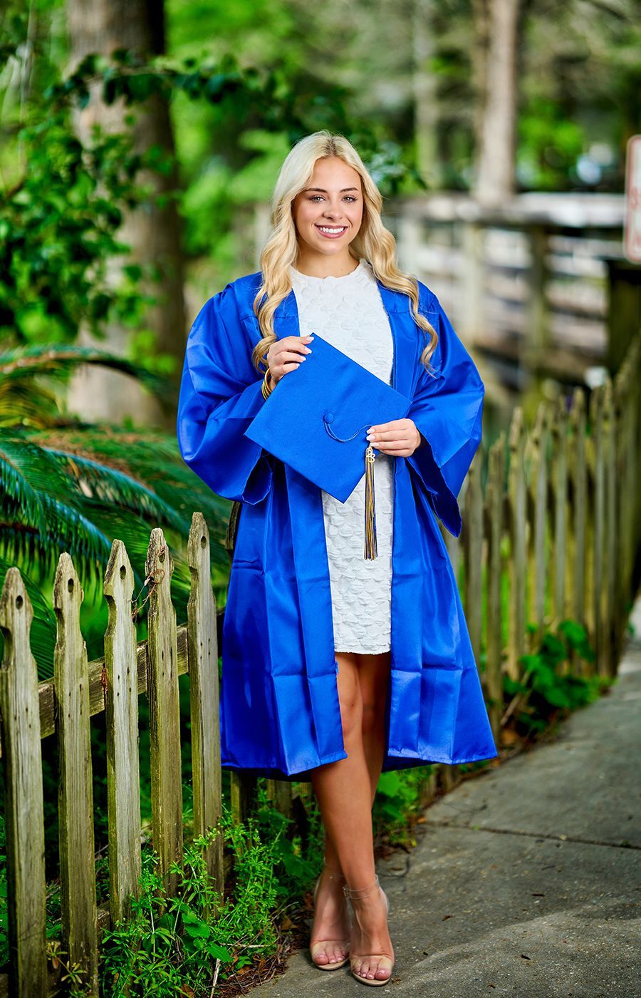 A woman in a blue graduation cap and gown is standing in front of a wooden fence.