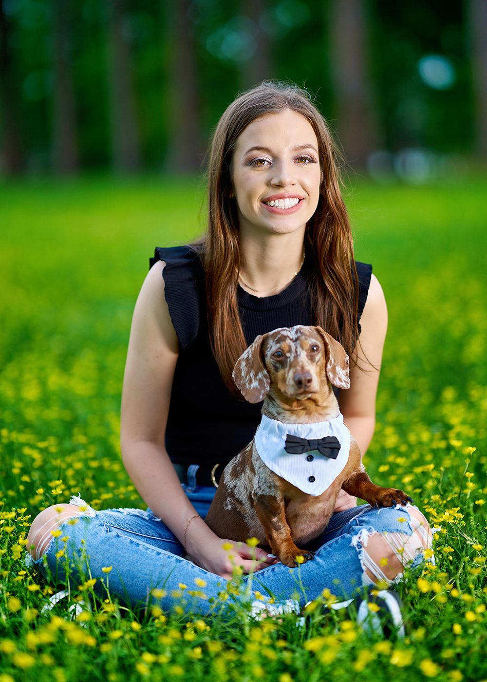 A woman is sitting in a field of flowers with a dachshund wearing a bow tie.