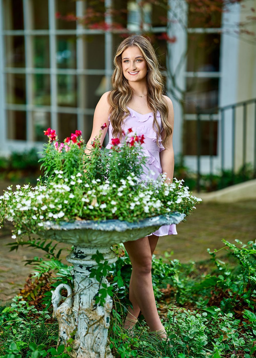A woman in a purple dress is standing next to a bird bath filled with flowers.