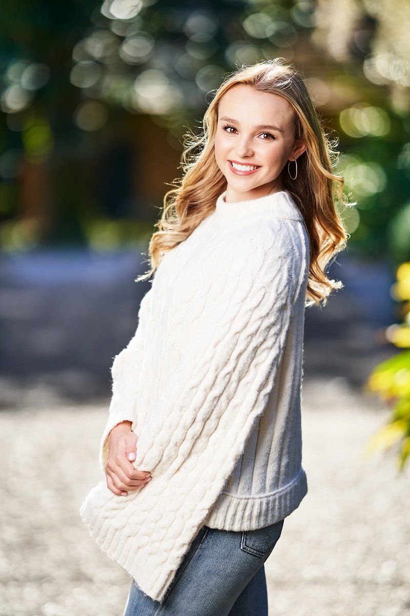 A young woman wearing a white sweater and jeans is standing on a sidewalk.