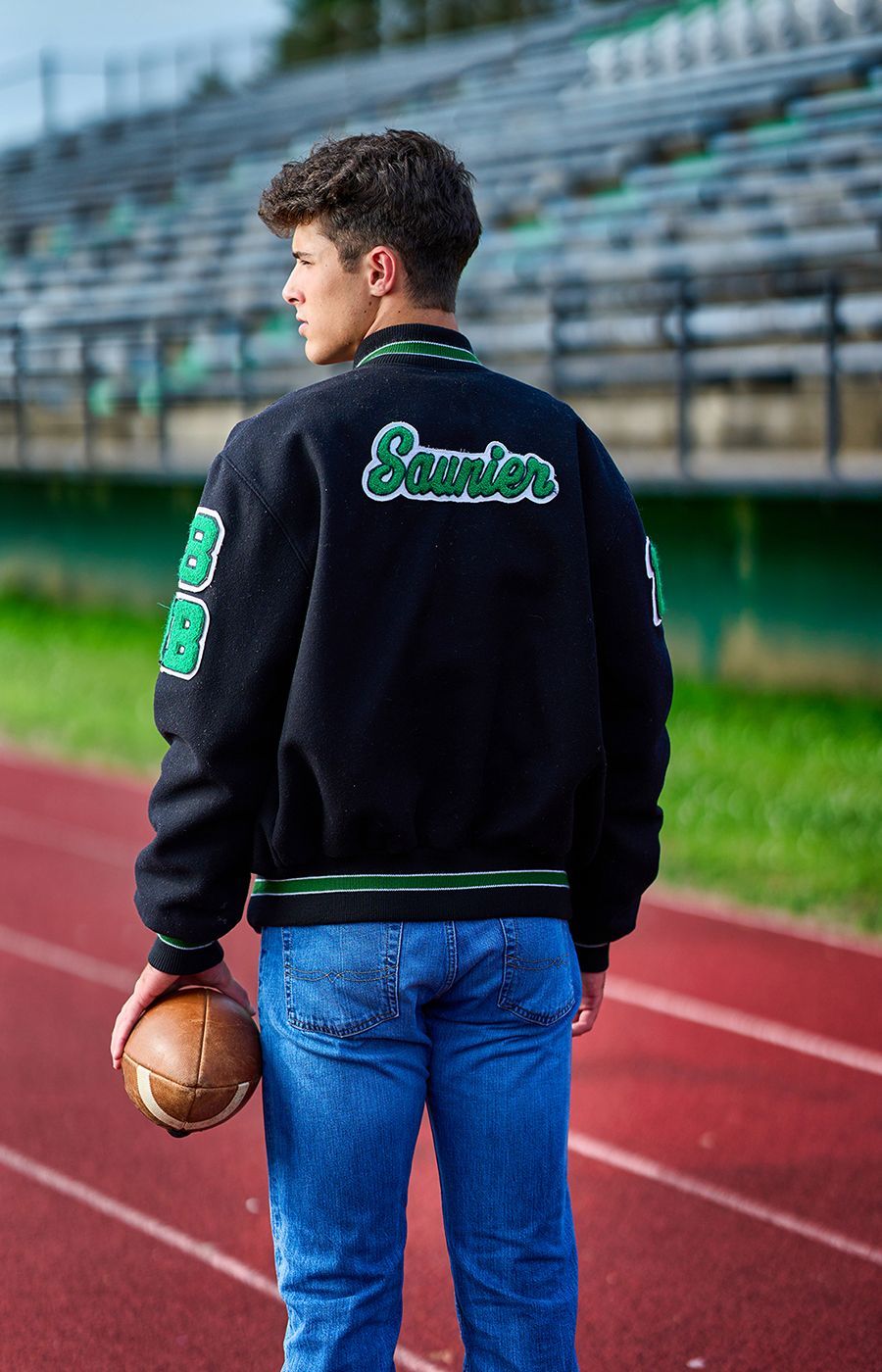 A young man in a letterman jacket is holding a football on a track.