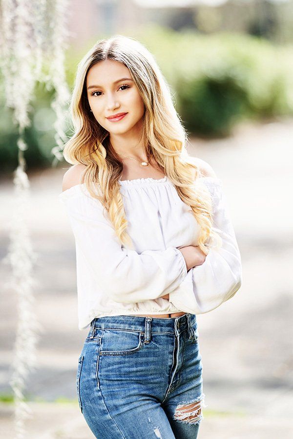 A woman in a white off the shoulder top and blue jeans is standing with her arms crossed.