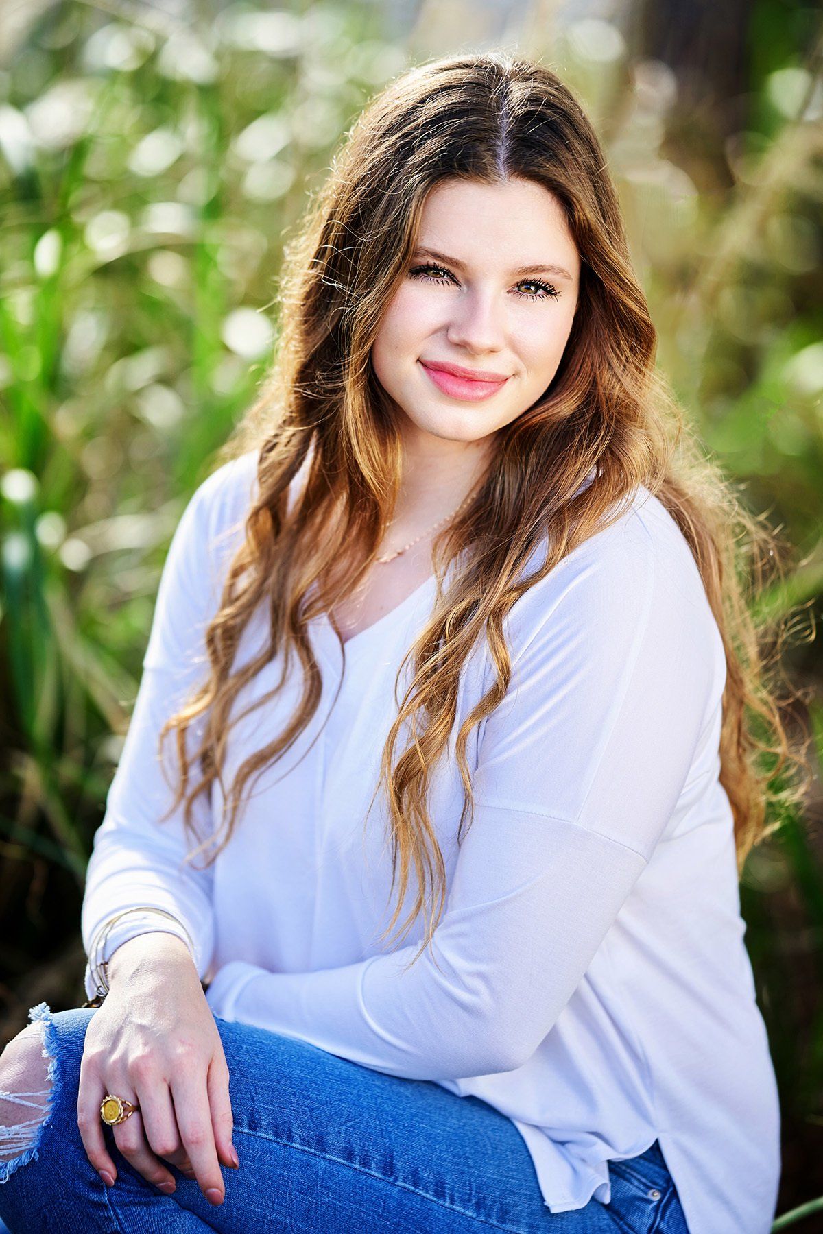 A young woman in a white shirt and blue jeans is sitting in the grass.