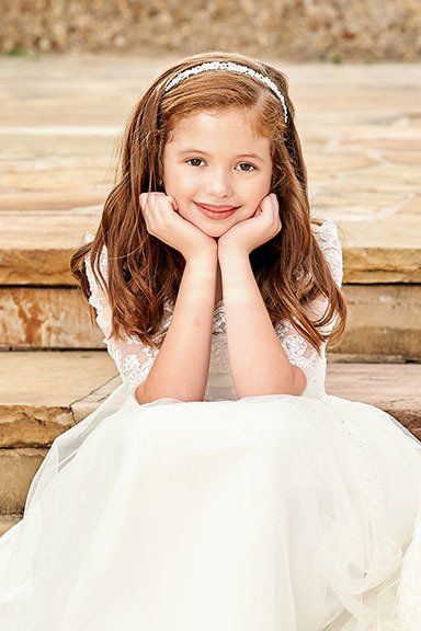 A little girl in a white dress is sitting on a wooden staircase with her hands on her chin.