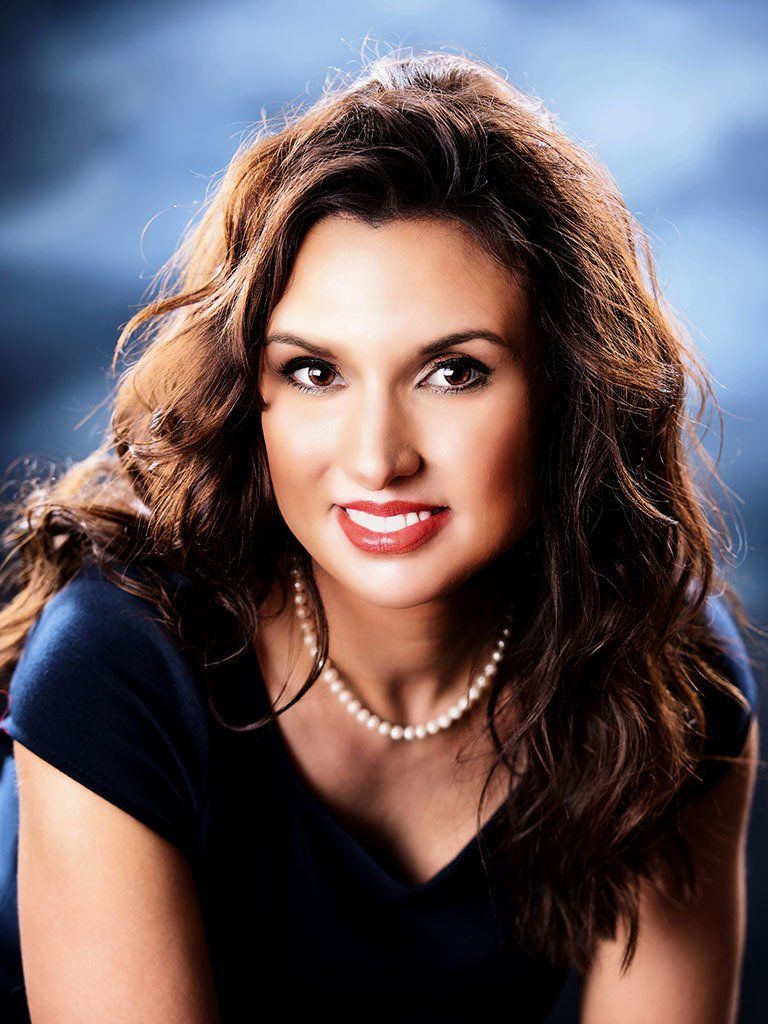 A woman wearing a pearl necklace and a blue shirt is smiling for the camera.