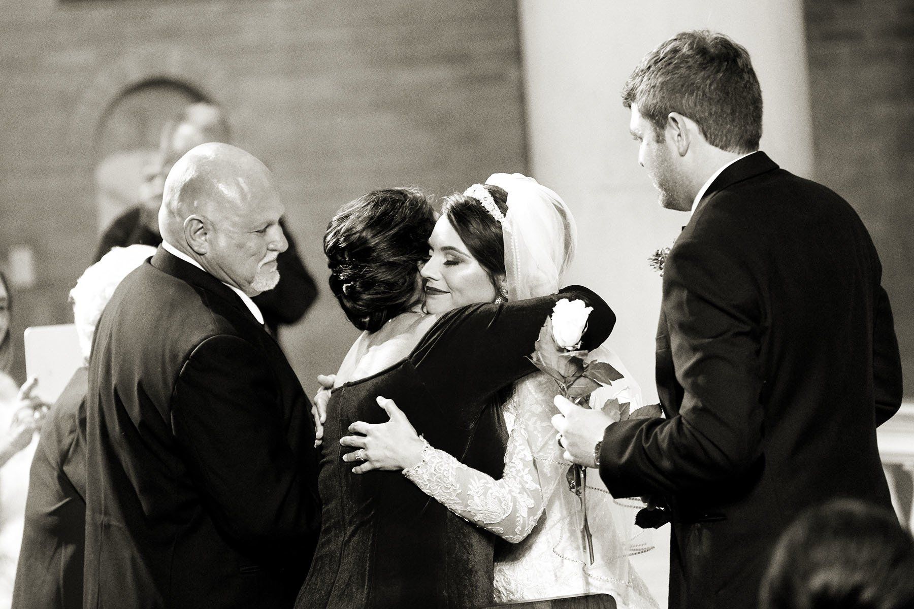 A bride and groom are hugging their parents at their wedding