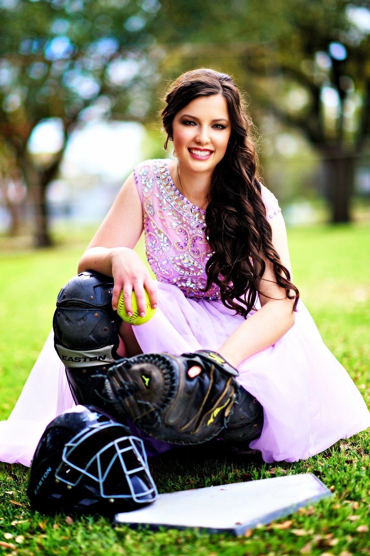 A woman in a pink dress is sitting on the grass with a baseball glove and helmet.