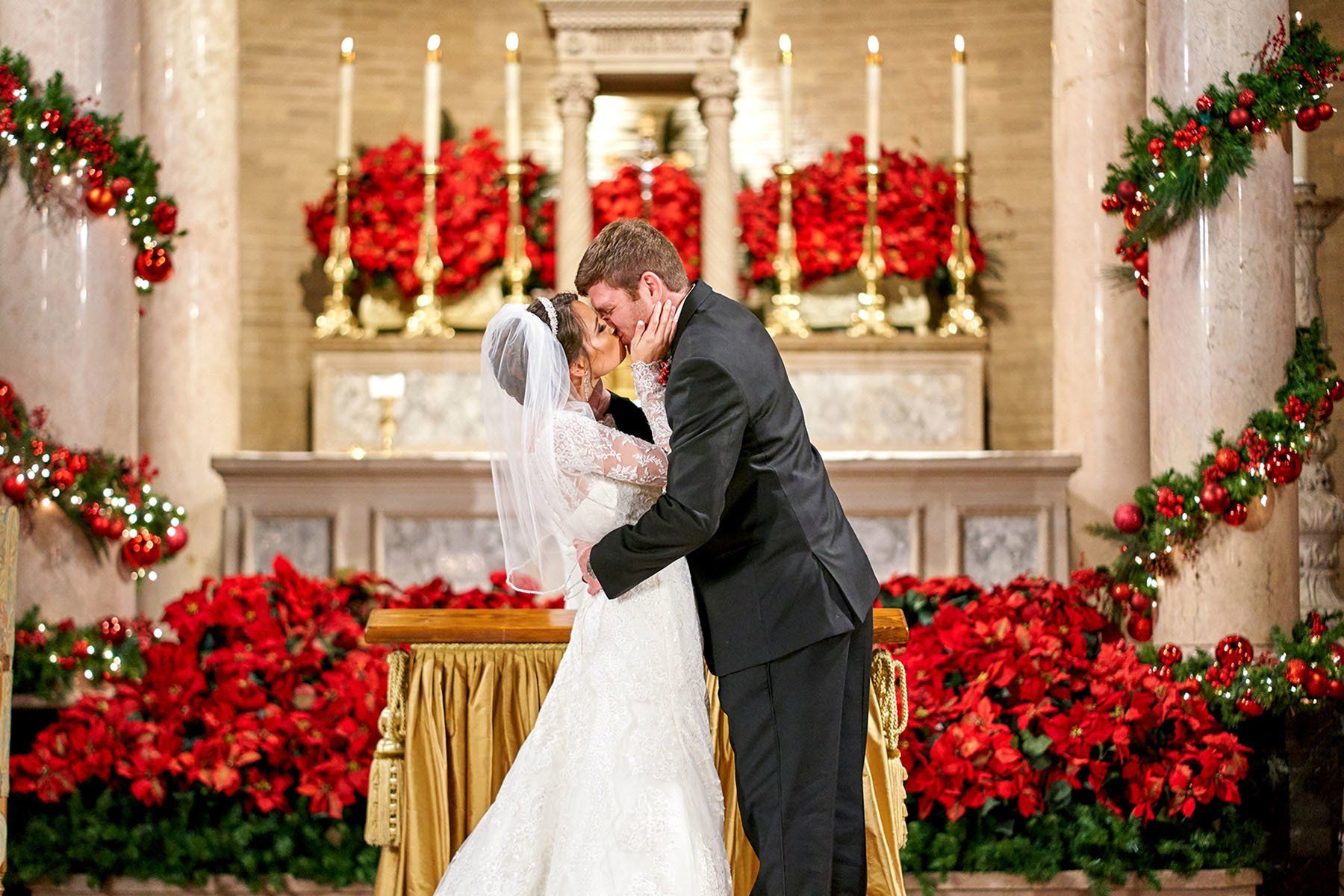 A bride and groom kissing in front of a church decorated with red flowers.