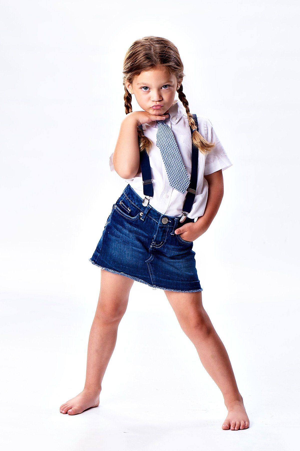 A little girl wearing suspenders and a tie is standing on a white background.