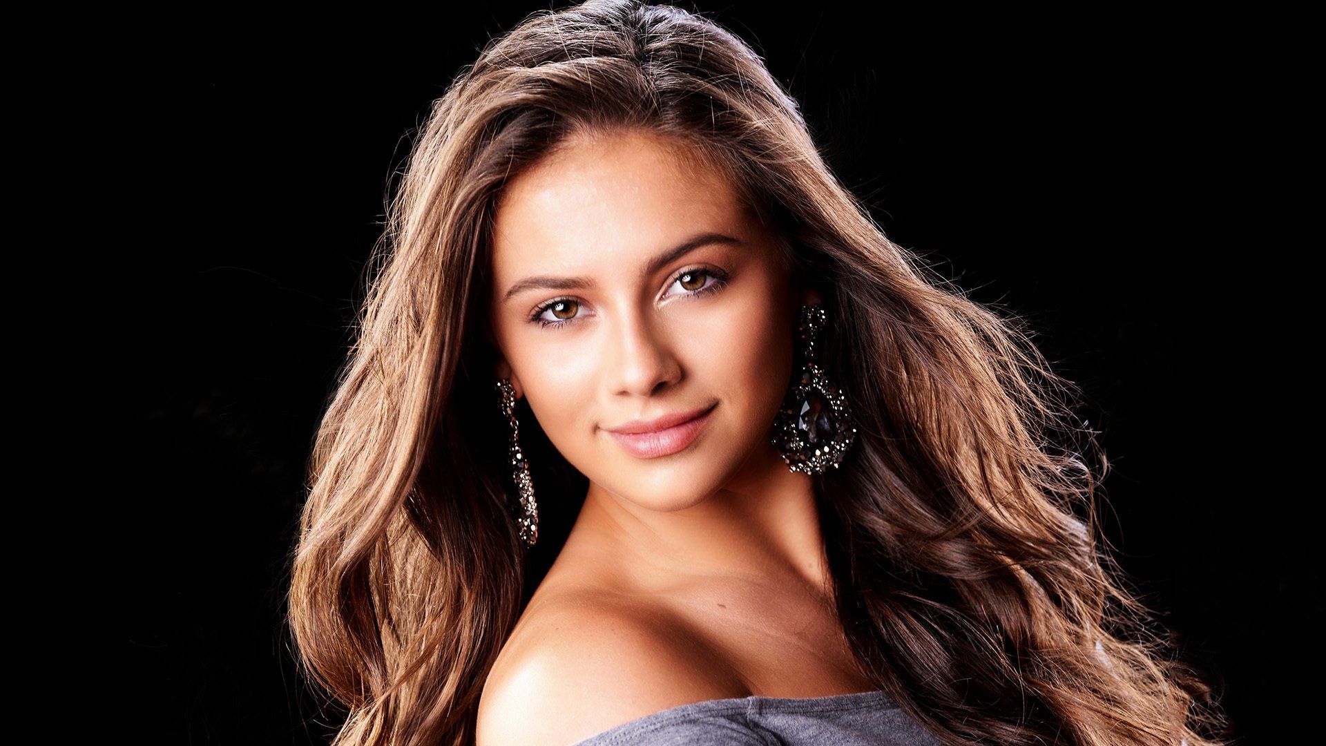A woman with long hair and earrings is standing in front of a black background.