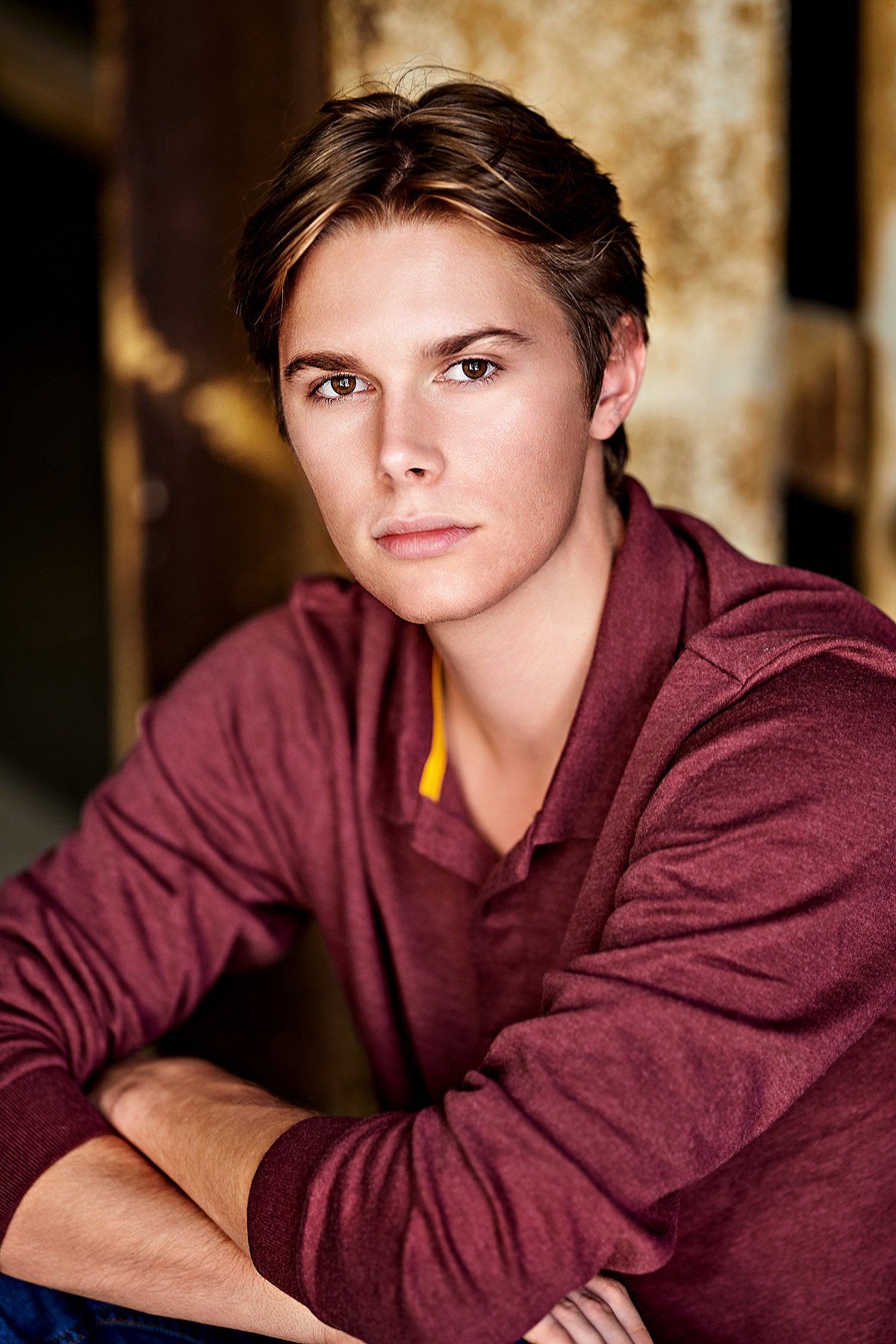 A young man in a maroon shirt is sitting down with his arms crossed and looking at the camera.