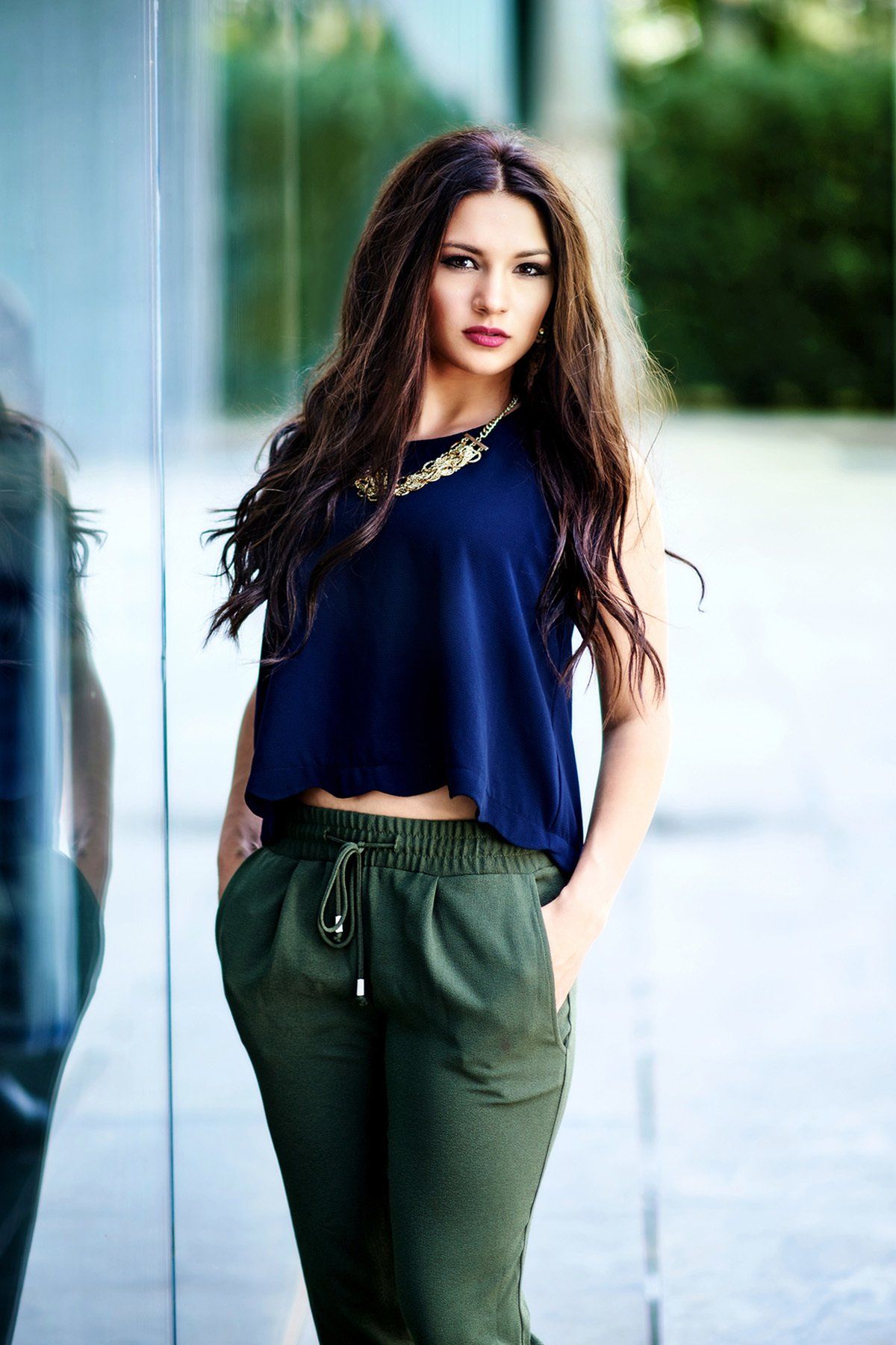 A woman in a blue crop top and green pants is standing in front of a glass wall.