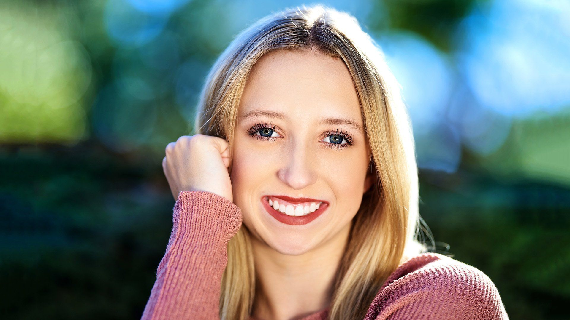 A woman in a pink sweater is smiling with her hand on her face.