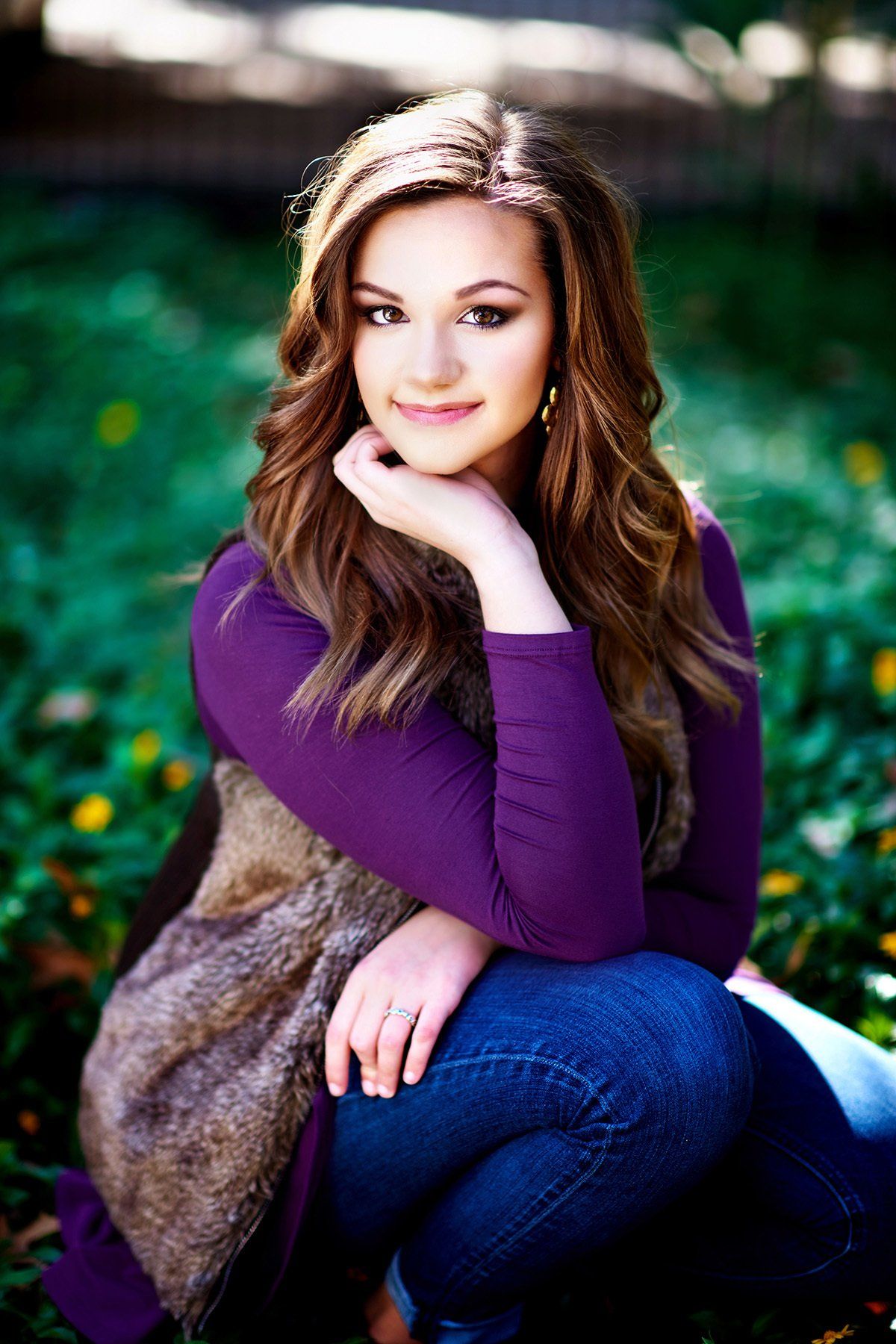 A woman in a purple sweater and fur vest is kneeling down in the grass.