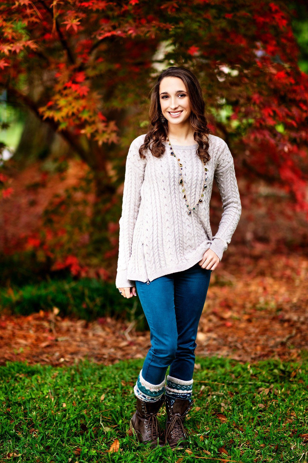 A woman in a sweater and jeans is standing in the grass in front of a tree.