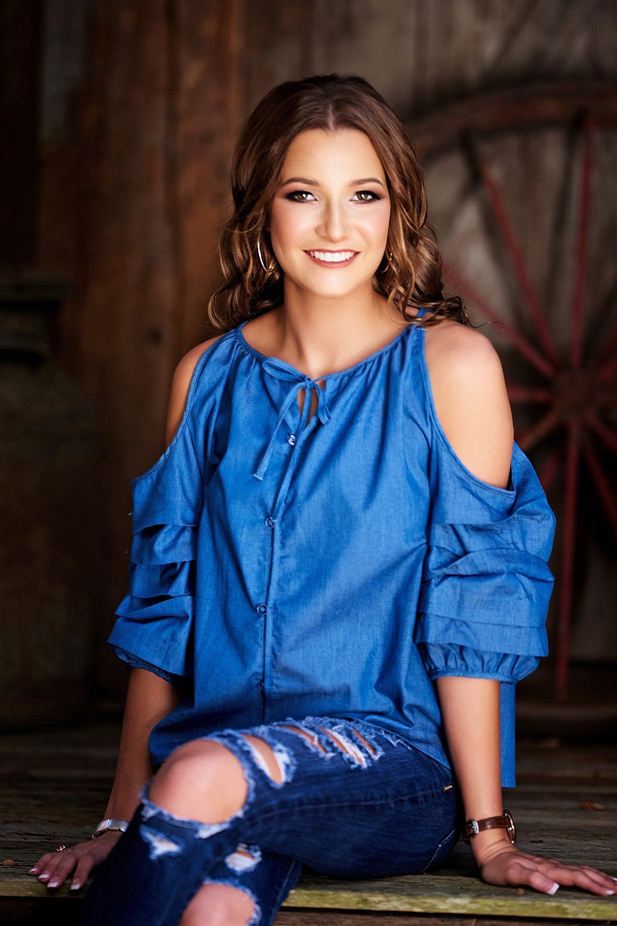 A woman in a blue off the shoulder top and ripped jeans is sitting on a bench.