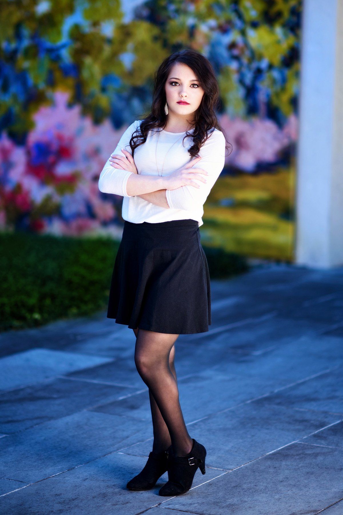 A woman in a white shirt and black skirt is standing on a sidewalk with her arms crossed.