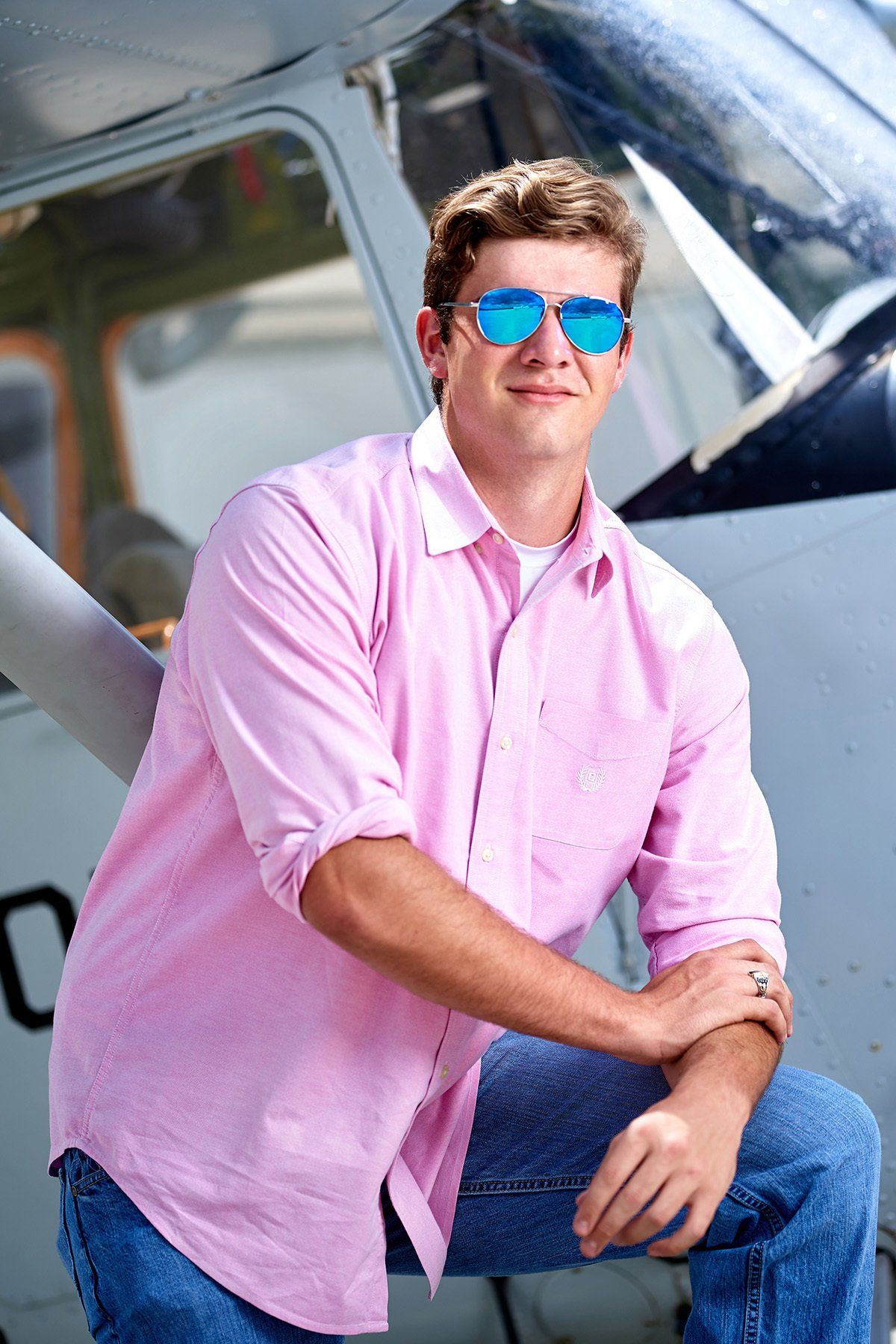 A young man wearing sunglasses and a pink shirt is sitting in front of an airplane.