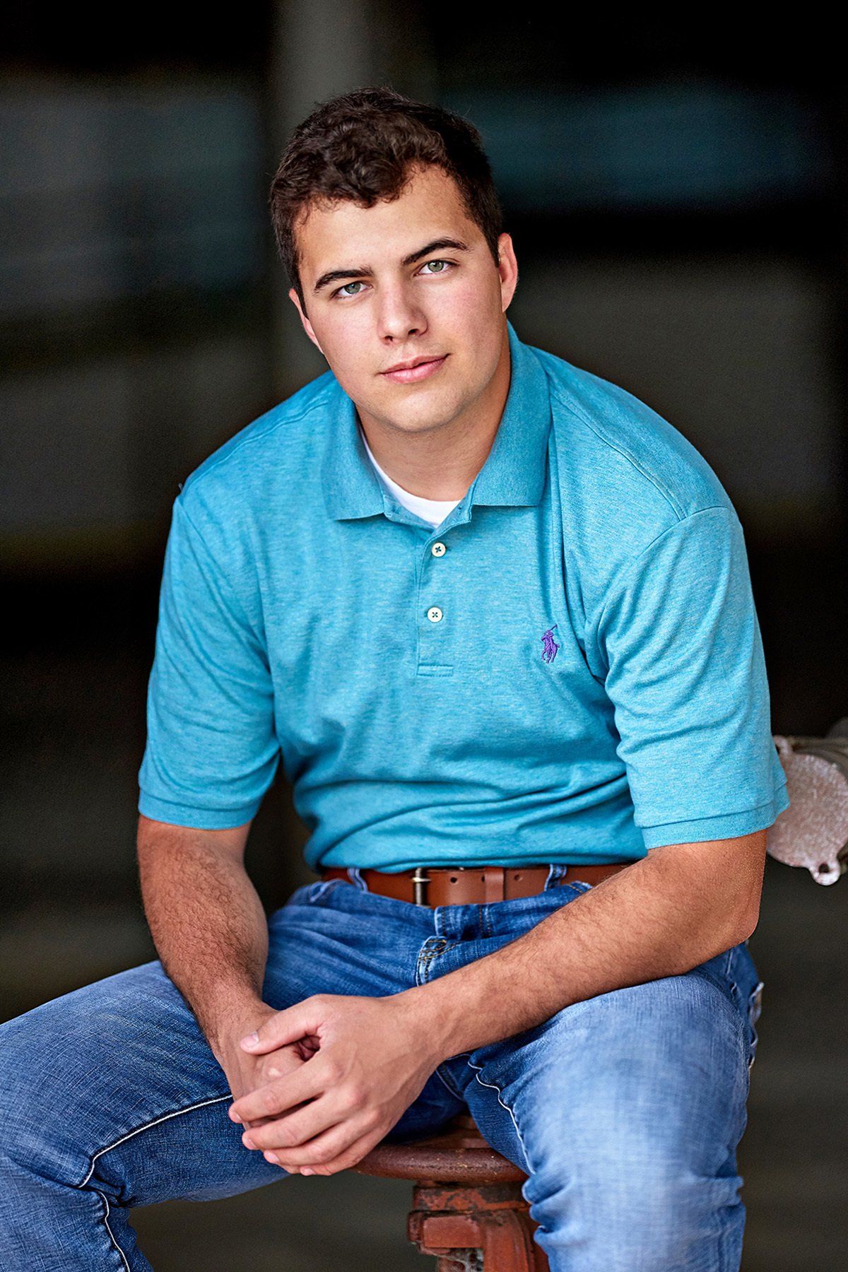 A young man in a blue polo shirt and jeans is sitting on a stool.