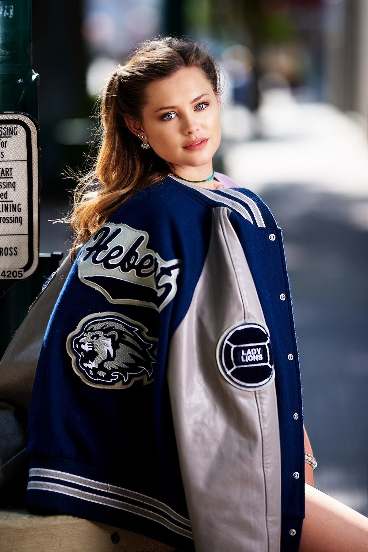 A woman wearing a blue and gray varsity jacket is sitting on a curb.