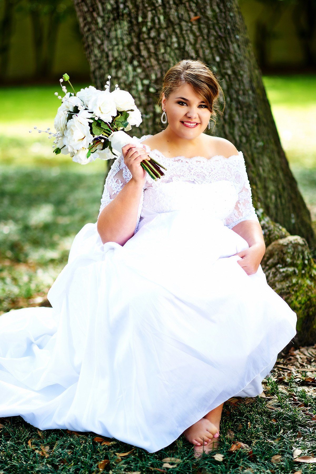 A woman in a wedding dress is sitting under a tree holding a bouquet of flowers.