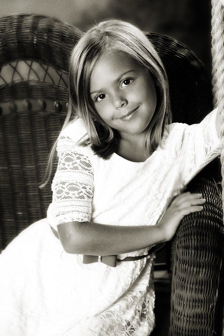 A little girl in a white dress is sitting in a wicker chair
