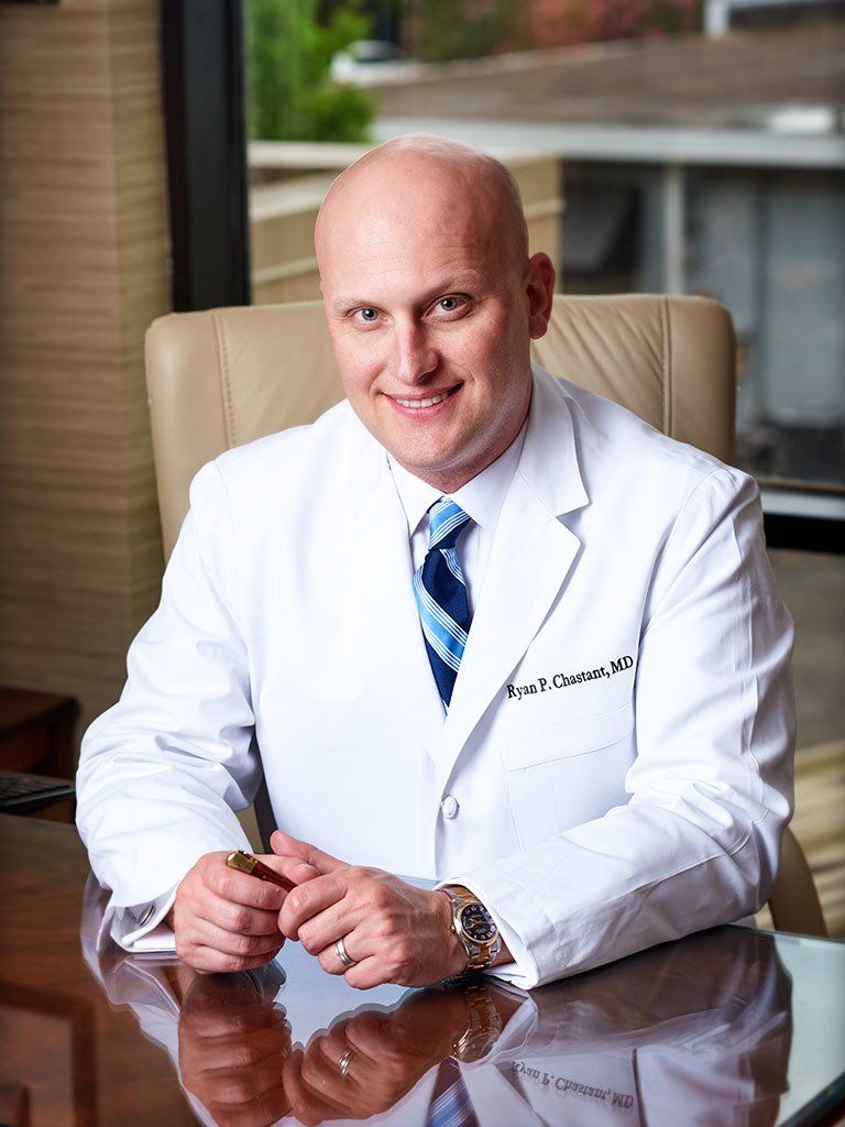 A bald man in a white coat and tie is sitting at a desk.