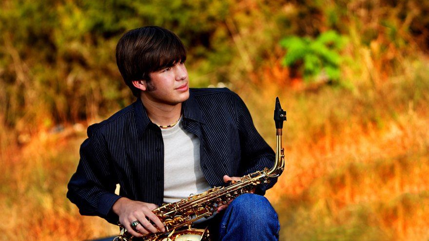 A young man is sitting in a field holding a saxophone.
